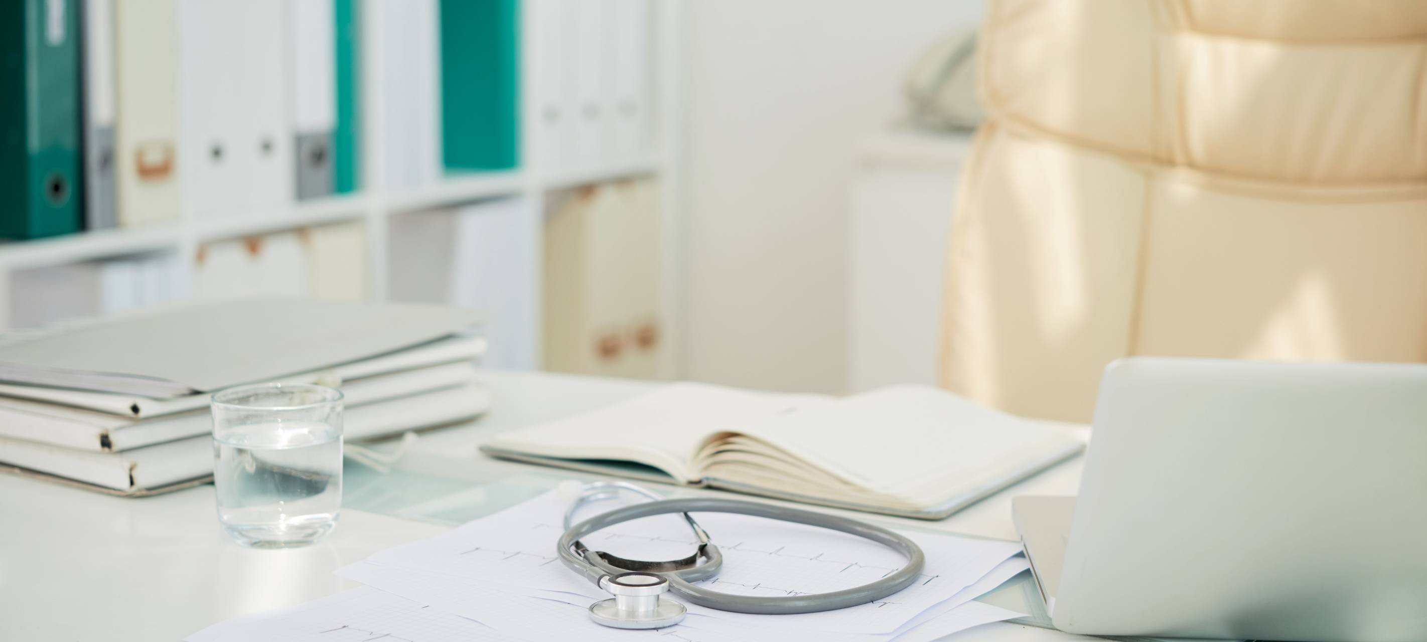 Schreibtisch in einer Arztpraxis auf dem Unterlagen, ein Laptop und ein Stethoskop liegen.